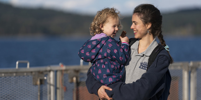 a young woman in a grey polo and navy sweatshirt holds a toddler girl with blond hair in front of a body of water.
