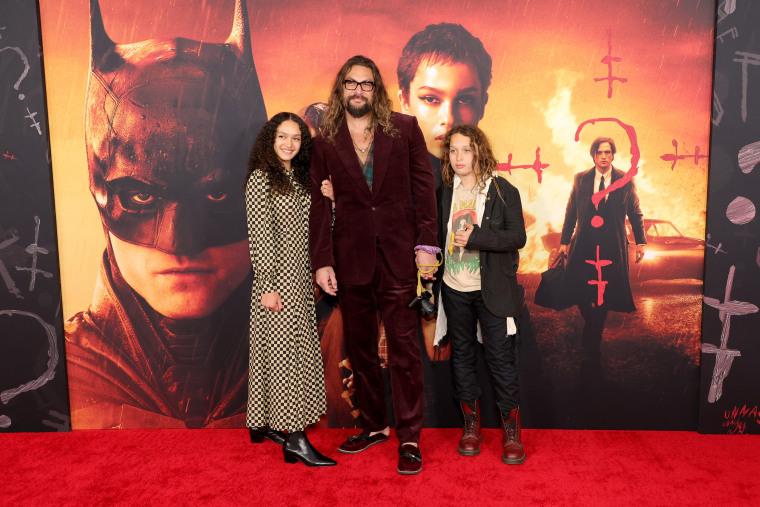 Mamoa in a purple velvet suit with his two young kids standing in front of a batman poster on the red carpet
