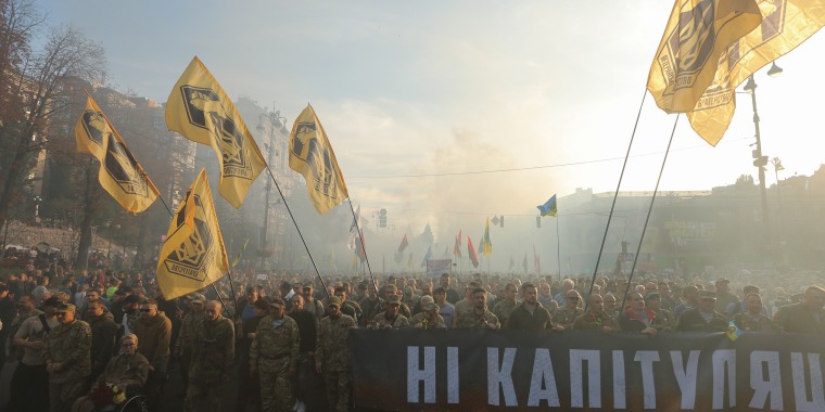 Image: People in the uniform carrying Azov battalion flags and a banner saying \"No Capitulation\" through the smoke during a rally in Kyiv, Ukraine, Oct. 14, 2019.