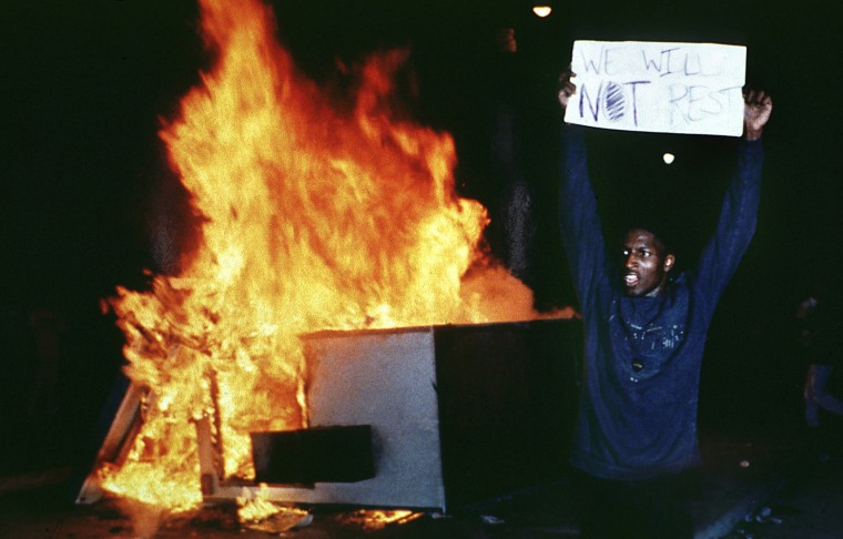 Image:A man holds up a sign that reads,\"We will not rest\" in front of a fire.