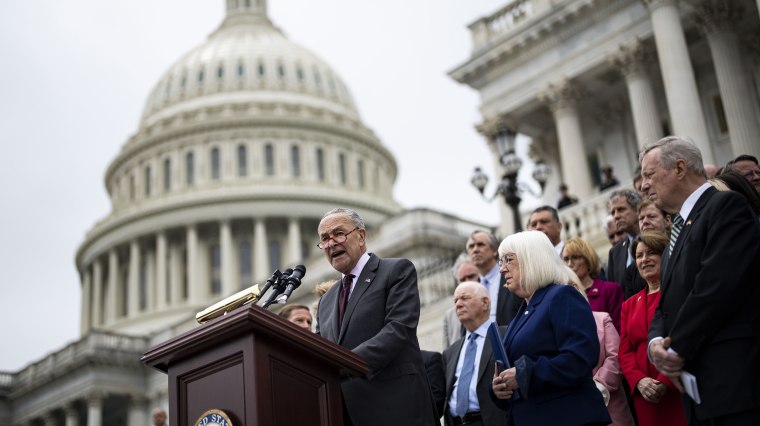 Image: Chuck Schumer speaks on the podium with the Senate Democrats standing behind him on the steps of the U.S. Capitol.
