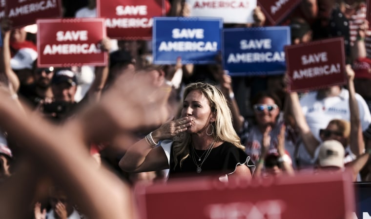 Image: Marjorie Taylor Greene on a stage at a rally with people in the crowd holding signs that read,\"Save America\".