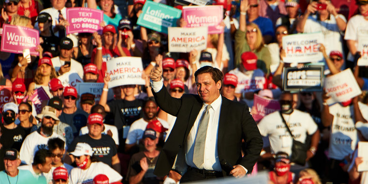 Image: Ron DeSantis speaks to a crowd during a campaign rally. A sign in the audience reads,\"Latinos for Trump\".
