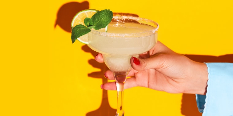 Female hand holding glass with margarita cocktail isolated on bright yellow neon background with shadow. Concept of taste, alcoholic drinks