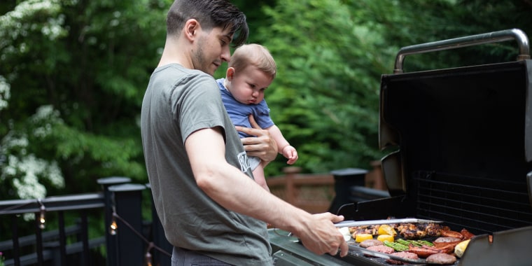 A man holding a baby, while grilling some meat and vegetables