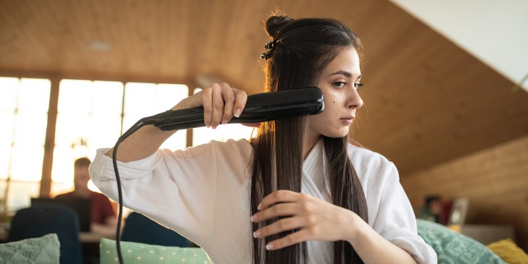 Young woman in bathrobe using hair straightener at home