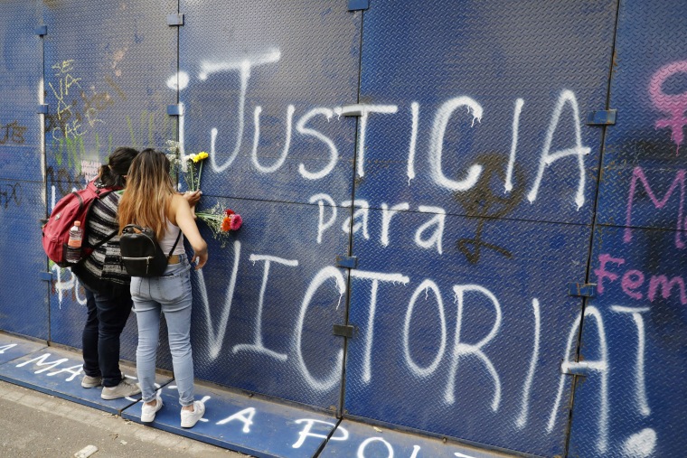 Young women place flowers on the perimeter wall of the Quintana Roo state offices sprayed with graffiti that reads in Spanish "Justice for Victoria," during a protest in Mexico City, Monday, March. 29, 2021.