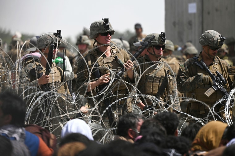 U.S. soldiers stand guard behind barbed wire as Afghans sit on a roadside near the military part of the airport in Kabul on Aug. 20, 2021.