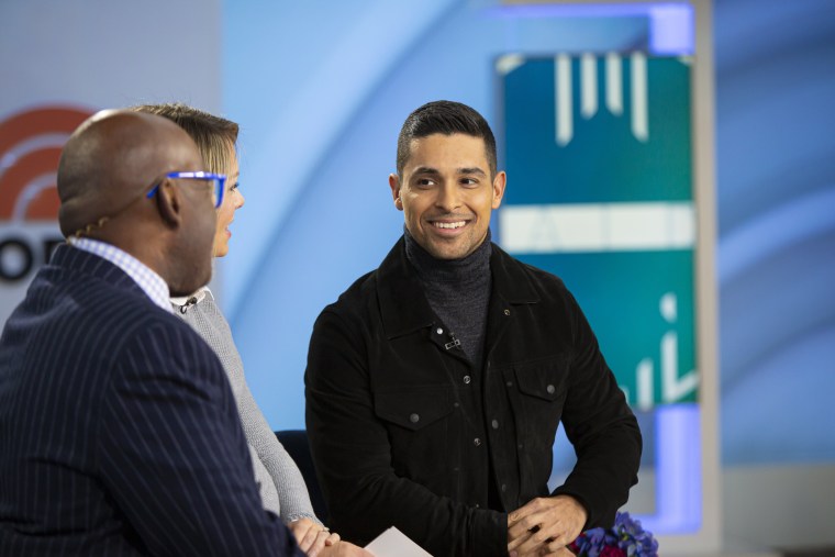 Valderrama in a black jacket and gray turtleneck sits on a TV interview set