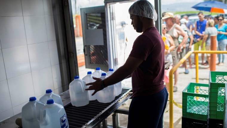 Un trabajador con gorra de redecilla junta botellas plásticas de agua en una planta de envasado de agua potable.