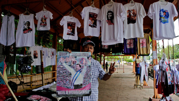 Un hombre sostiene un cartel con Daniel Ortega y la leyenda "Hasta la victoria siempre" enfrente de camisetas blancas colgadas que tienen diseños de Daniel Ortega, a modo de mercancía para una campaña electoral.