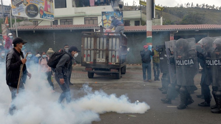 En una calle ecuatoriana, del lado izquierdo hay un grupo manifestantes rodeados por gas lacrimógeno y del lado derecho un grupo de policías antimotines avanzando con escudos durante una huelga en la provincia de Cotopaxi.