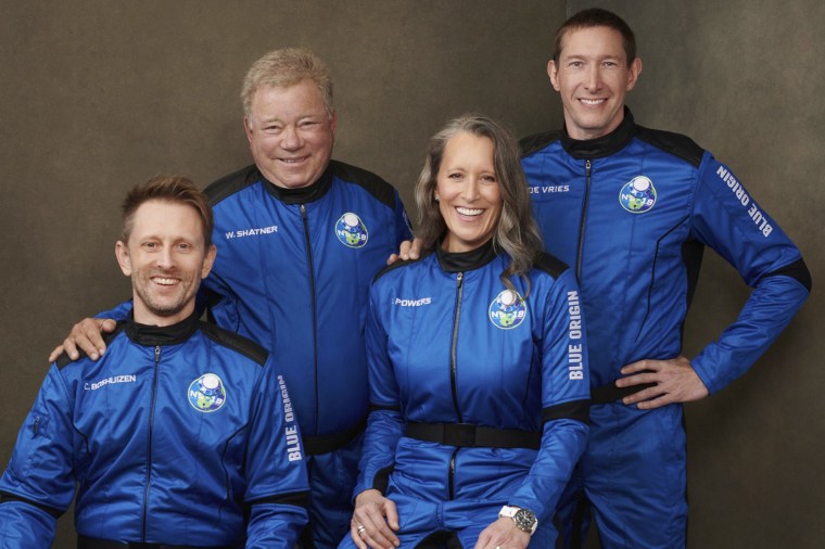 Three white men and one white woman in blue jumpsuits that read Blue Origin smile at the camera