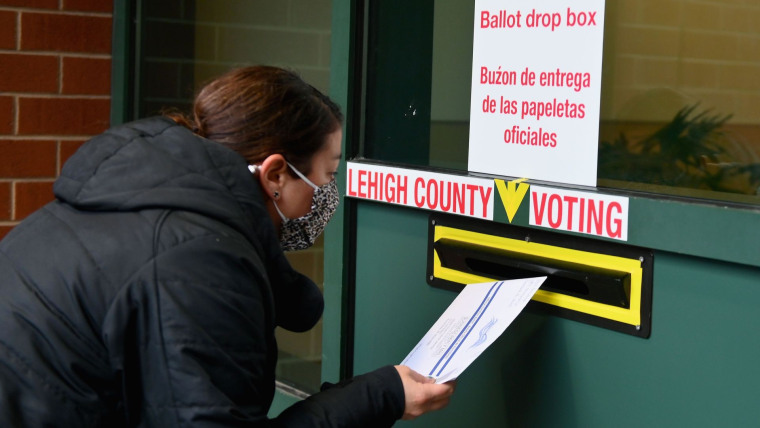 Una mujer con mascarilla y abrigo negro se inclina frente a un buzón electoral de color verde para depositar su boleta de voto anticipado en octubre de 2020 en Pennsylvania.