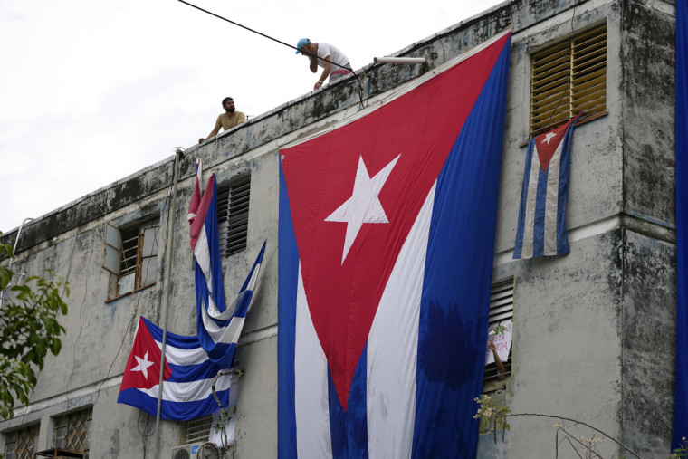 Hombres cuelgan banderas cubanas en las ventanas de la casa del dramaturgo y opositor Yunior García Aguilera en un intento para evitar que se comunique con el exterior, mientras éste sostiene una flor desde una ventana, en La Habana, Cuba, el domingo 14 de noviembre de 2021