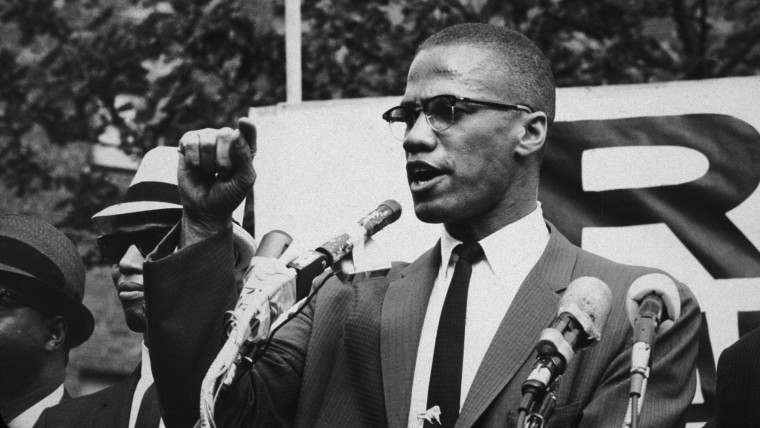 Malcolm X levanta el dedo índice derecho mientras da un discurso al aire libre, con varios micrófonos al frente
