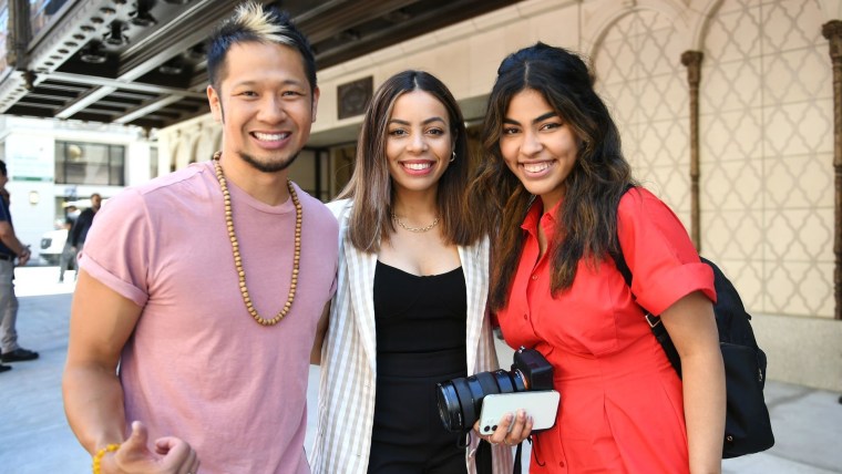 Tres personas sonríen juntas: de izquierda a derecha un hombre de ascendencia asiática con camisa color rosa, seguido de una mujer latina con saco de cuadros, y a la derecha otra mujer latina con una camiseta roja y sosteniendo una cámara fotográfica.