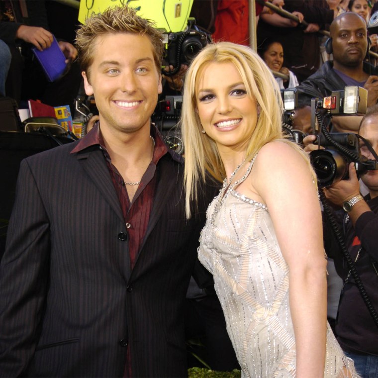 Lance Bass and Britney Spears smile on a red carpet. Bass is in a black suit with a red button-down shirt and Spears is in a sparkly, white gown