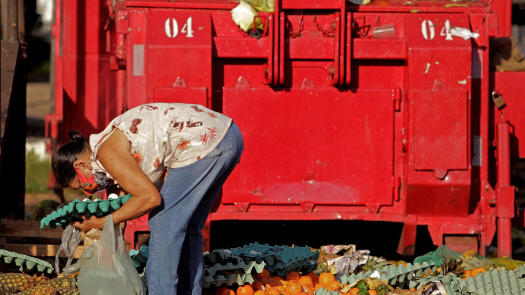 Una mujer brasileña con cubrebocas está agachada para escarbar entre la basura, como cajas de huevos y restos de fruta, de un mercado callejero en busca de comida.