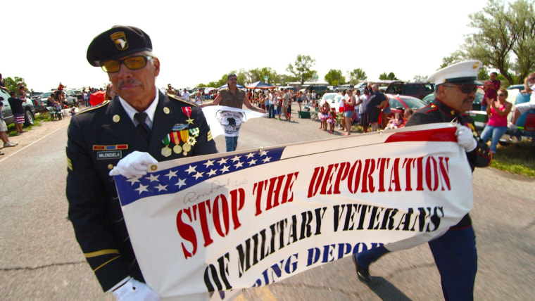 Dos hombres de edad avanzada vestidos con uniforme militar caminan por la calle durante un desfile cargando un cartel que dice "Detengan la deportación de veteranos militares".