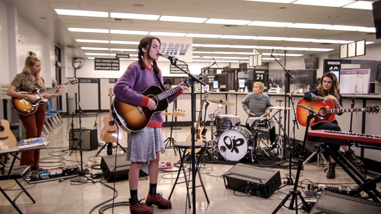 Olivia Rodrigo in a purple sweater and patterned skirt performs in an office space.