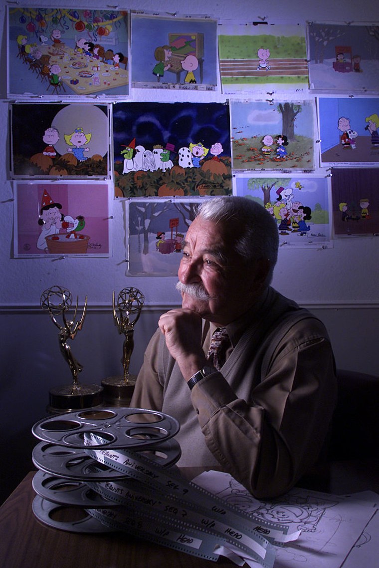Un hombre latino de edad avanzada con bigote posa enfrente de premios Emmy y de imágenes de las caricaturas de Snoopy y Charlie Brown