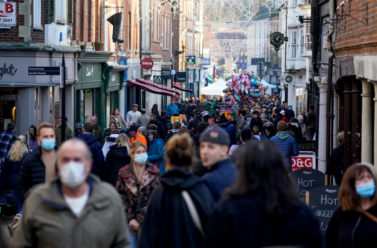 shoppers in winter clothes make their way down a crowded street. Some are in masks, others are not.
