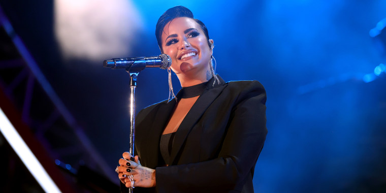 Demi Lovato in a black suit jacket smiles onstage holding a microphone.