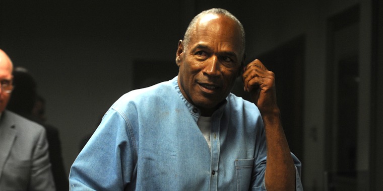 O.J. Simpson smiles as he looks off-camera with his left hand on his ear. he's wearing a light colored jean button down