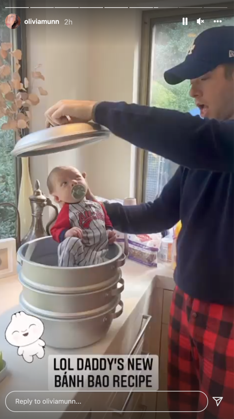 John Mulaney in a baseball cap and pajams holds a lid over his infant son, who is sitting in a pot on a countertop.