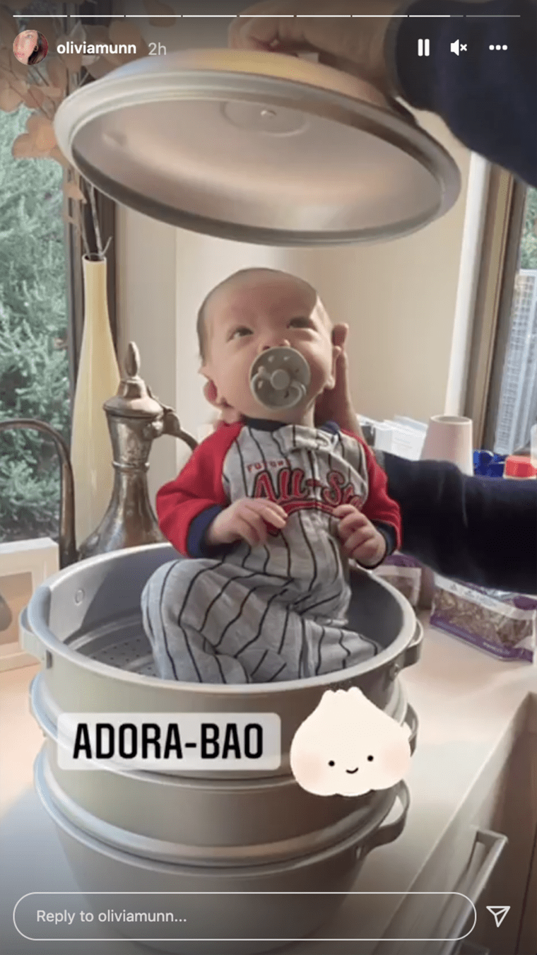 A young baby with a pacifier in a baseball-themed onesie sits inside a pot as a disembodied hand holds the lid over the baby.
