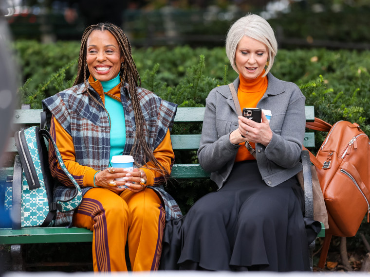 In trendy fall clothing, Cynthia Nixon and Karen Pittman sit on a green park bench holding takeout coffee cups. Nixon looks at a cell phone while Pittman smiles at something off camera.