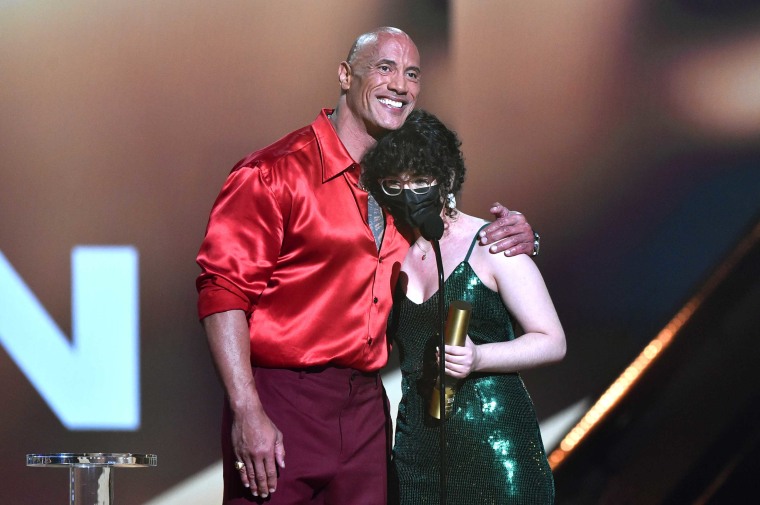 Johnson in a red silky shirt puts his left arm around a teen in a green sparkly dress as she holds his award in her left hand.