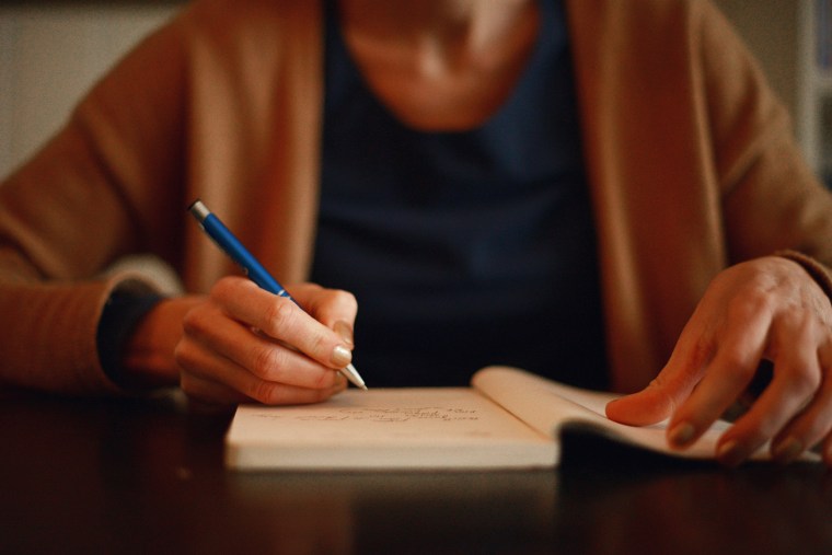 A person wearing a camel-colored sweater writes in a notebook.