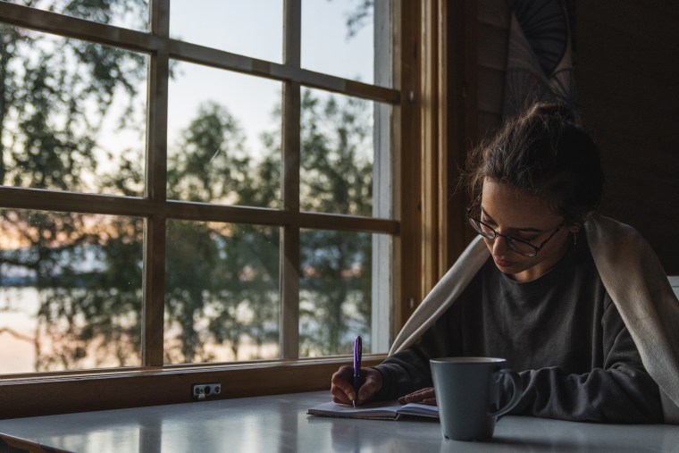 A woman sits by a window and writes in a journal while covering herself with a blanket.