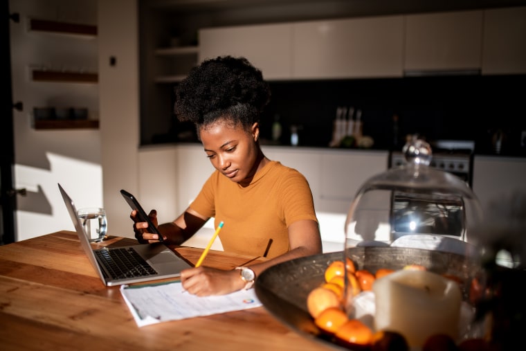 A woman holds a cellphone by a laptop while writing on a notepad. 