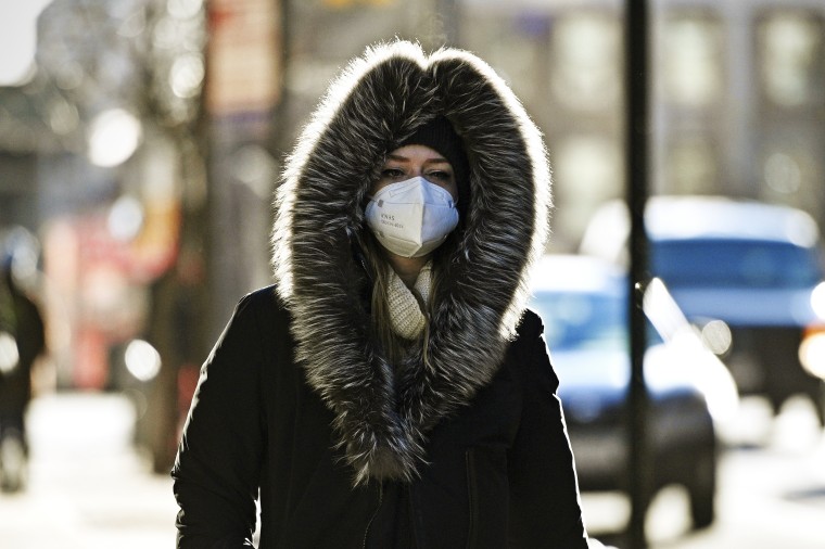 A woman wears a white medical-grade KN95 mask outside. She has her heavy overcoat on and the fur-lined hood pulled up around her head. She walks on a busy New York City street.