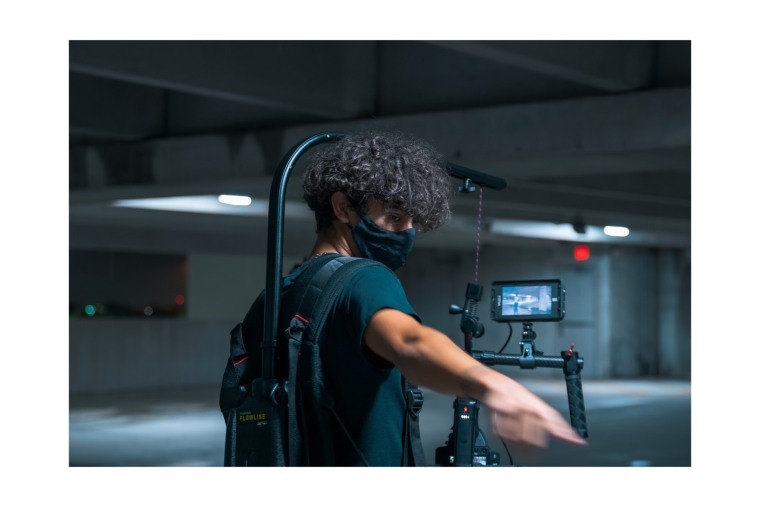 Un joven latino de cabello rizado sostiene una cámara y micrófono mientras dirige un cortometraje.