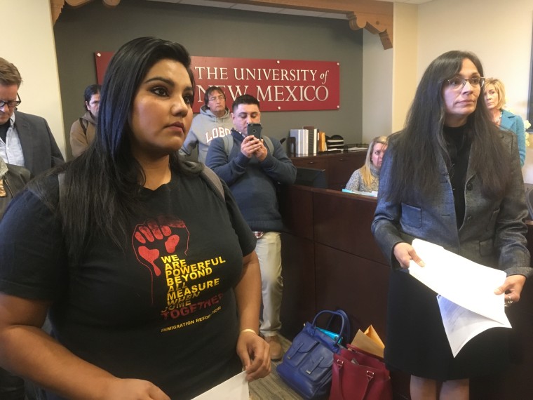 Una joven estudiante latina con camiseta negra escucha a alguien fuera de la toma al lado de una mujer con saco gris y lentes dentro de la Universidad de Nuevo México.