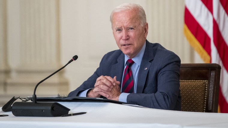 Joseph Biden, cabello canoso, con traje azul marino y corbata roja, entrelaza las manos enfrente de un micrófono
