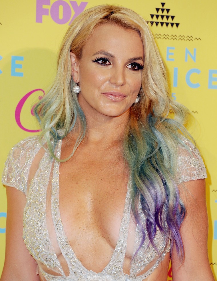 Spears smiles in front of a yellow wall. She has blond hair with green and purple ends styled in waves. She's wearing a sparkly gold dress.