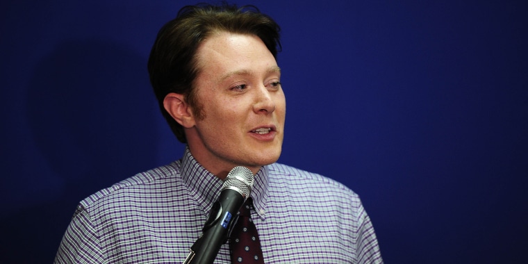 Clay Aiken with brown hair stands in front of a mic, speaking, in a plaid button-down shirt and tie.
