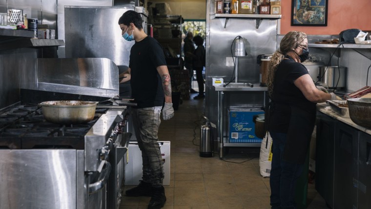 Un chef latino joven está parado enfrente de la estufa de un restaurante del lado izquierdo y del lado derecho de la misma cocina una mujer latina prepara alimentos sobre una barra