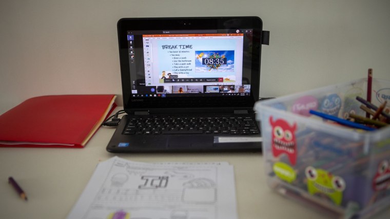 Una laptop pequeña con un programa educativo yace sobre un escritorio junto a un cuaderno rojo y una caja de lápices.
