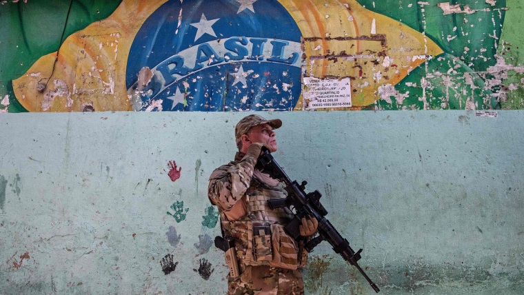 Un hombre en vestimenta militar y con un arma larga en mano se para enfrente de un mural desgastado con la bandera de Brasil y manos pintadas por niños.