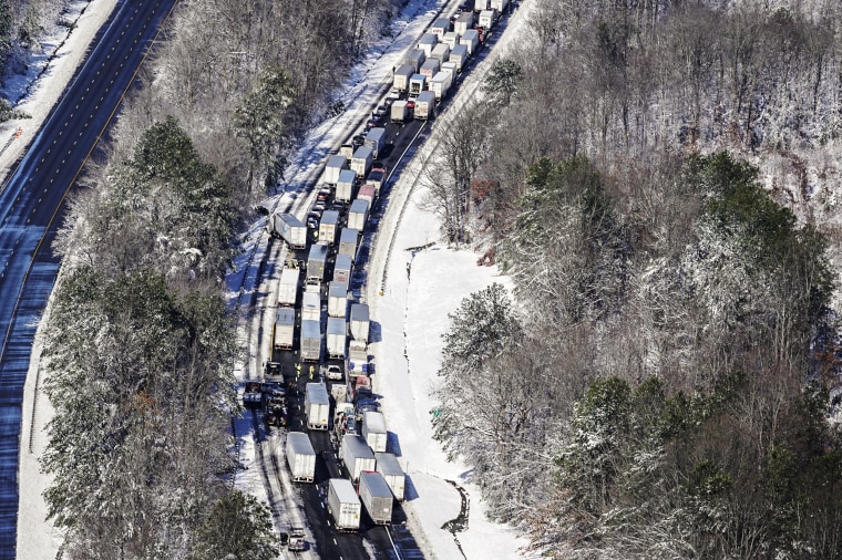 Cars and trucks sit completely stopped on a highway in a snowy forest. One truck appears to be stuck slightly off the road.