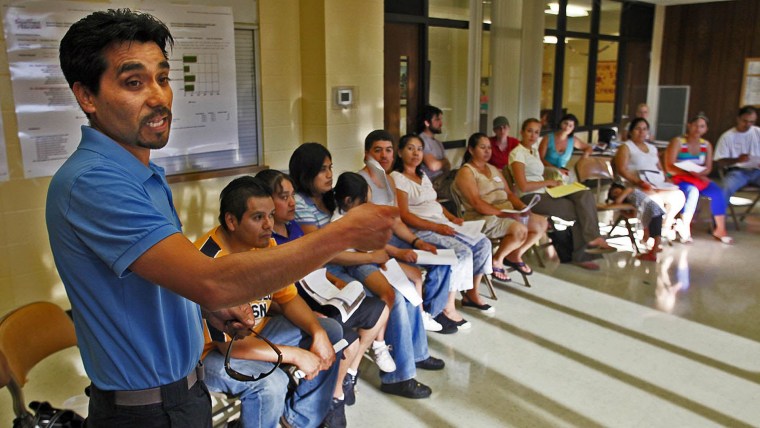 Un hombre latino de camisa azul gesticula mientras habla en una reunión de primaria con otros padres de familia, sentados y con caras serias