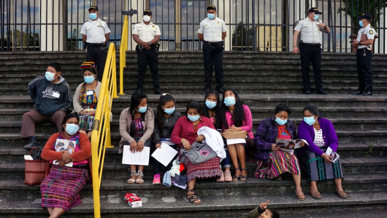 Nueve mujeres y niñas guatemaltecas con indumentaria tradicional maya se sientan en escaleras grises con papales en sus manos