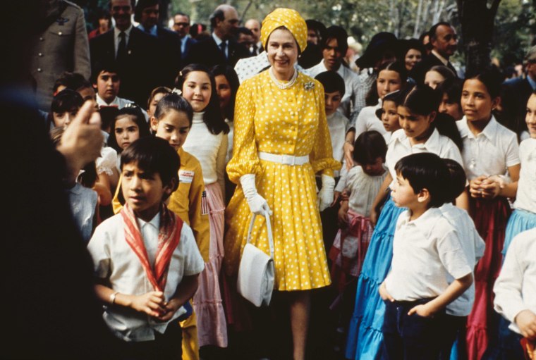 The Queen wears a yellow polka dotted a line dress and coordinated turban as she stands among a group of schoolchildren.