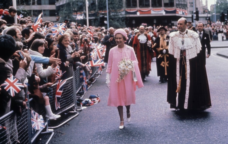 Queen stands in pink dress, jacket and hat on a street next to a man in a floor length robe with white fur. Cheering crowds holding british flags behind barricades wave.